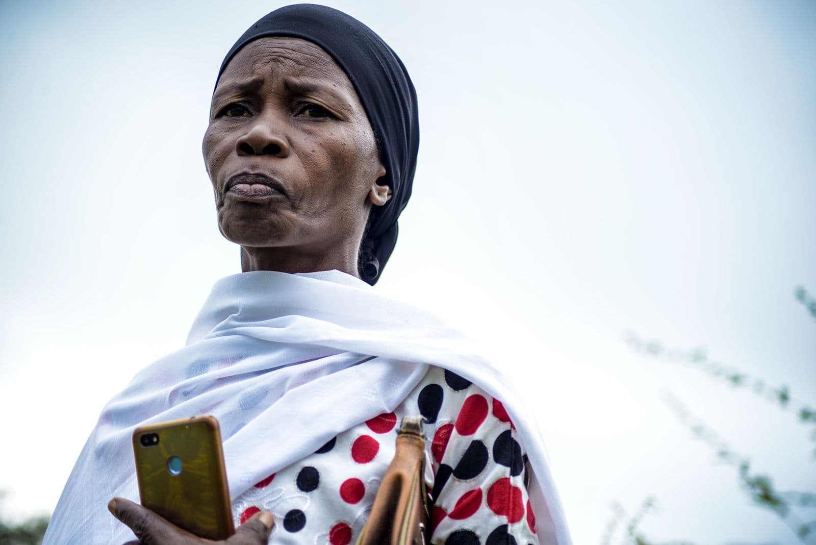 A Tanzanian woman in traditional dress holding a smartphone outdoors, the kind of phone used to invest in UTT AMIS via M-Pesa.
