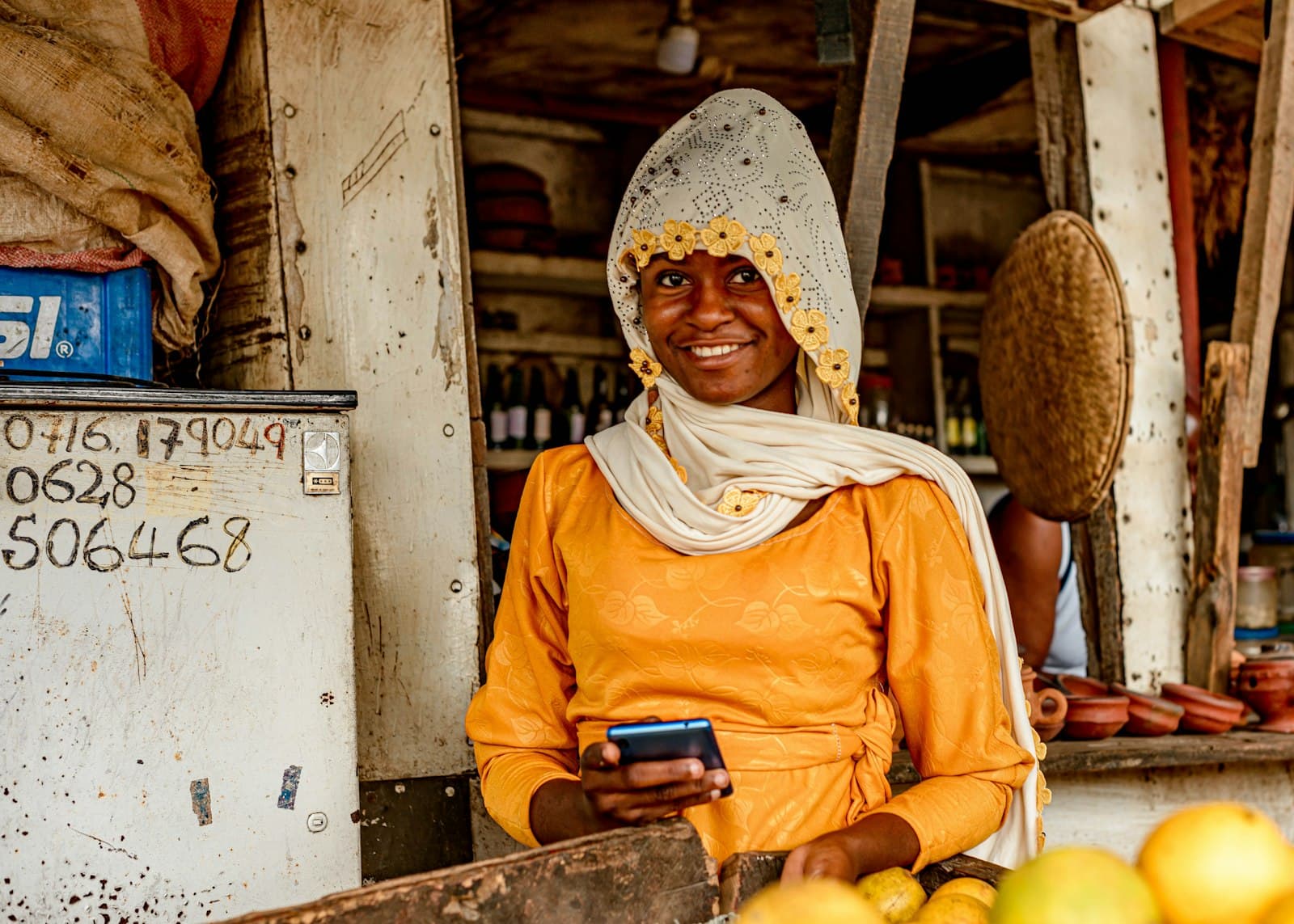 A woman at a market stall in Tanzania holding her phone, the kind of everyday spot where M-Pesa Tanzania payments happen