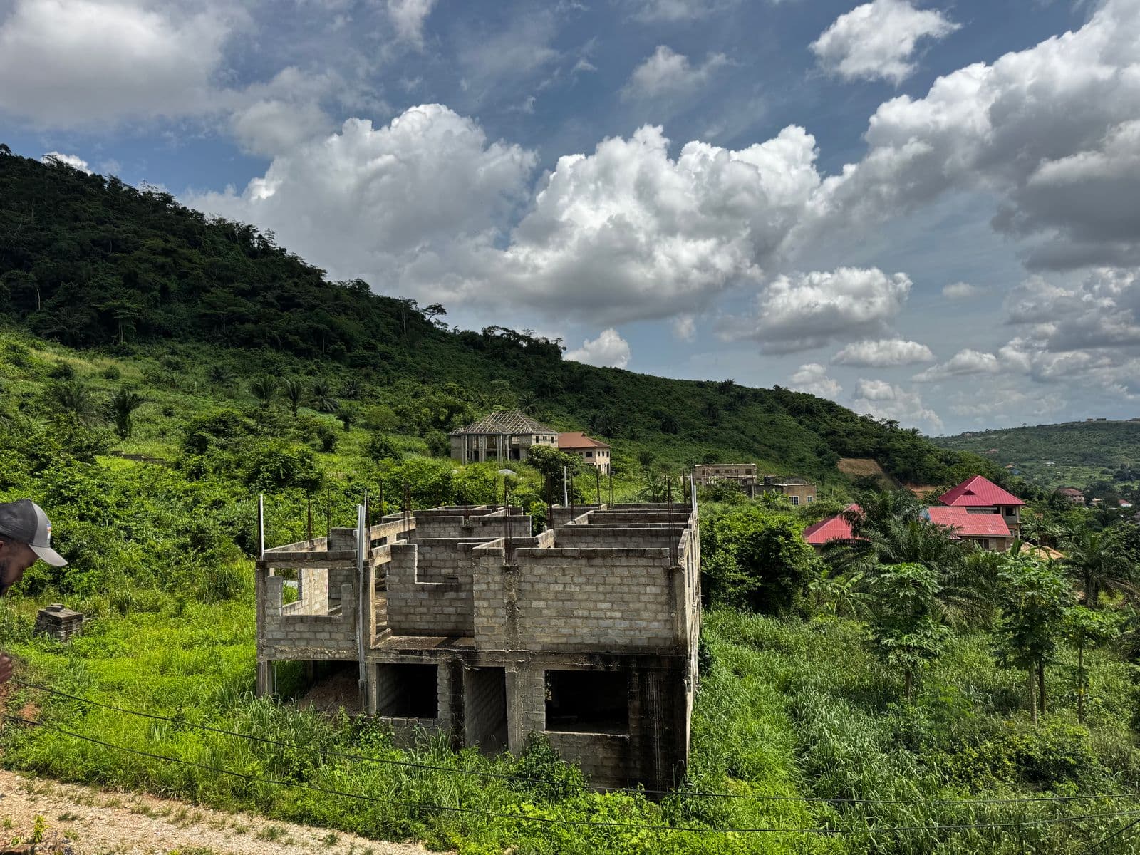 An unfinished house mid-construction in Greater Accra, Ghana, with block walls up and greenery around the site.