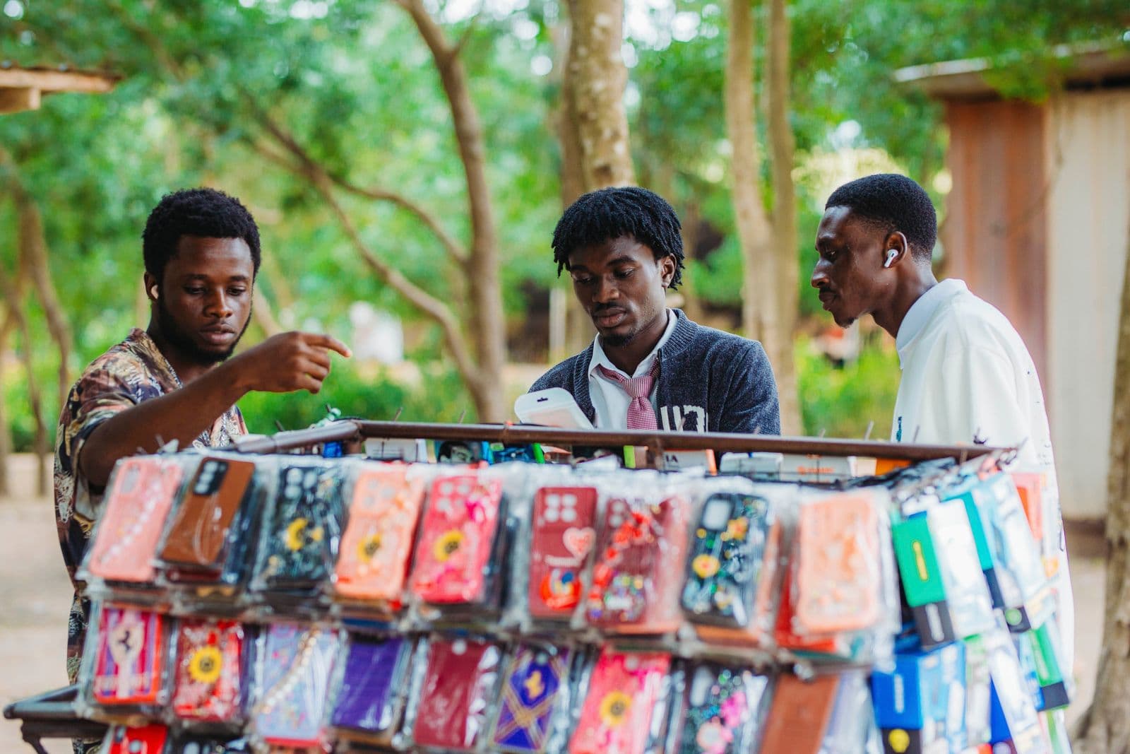 Three men browsing phone cases at an outdoor market stall in Accra, Ghana
